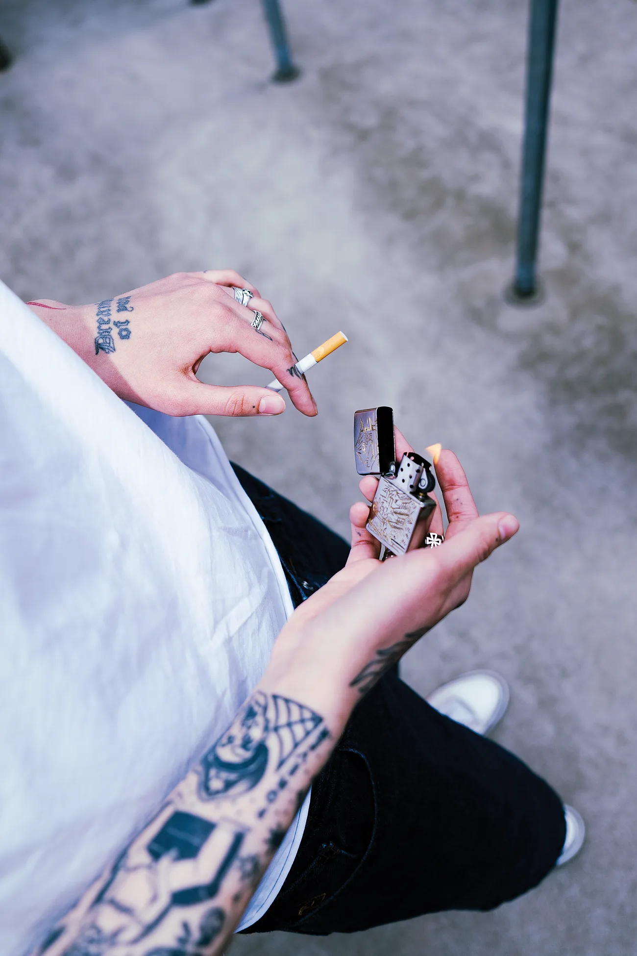 Tatuiruotos rankos su sidabriniais žiedais virš graviruoto Zippo – kadras iš viršaus.