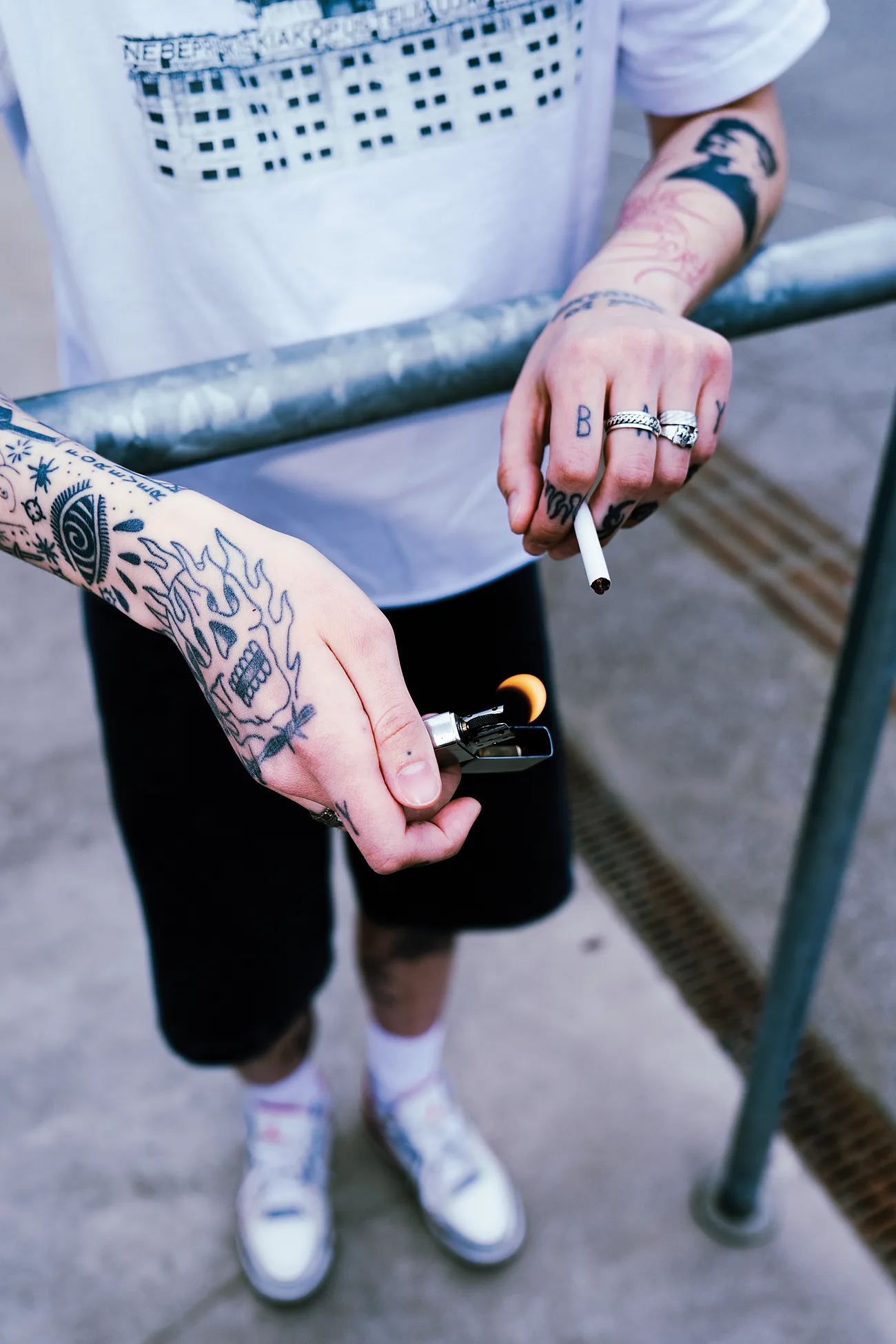 Tatuiruotos rankos prie plieninio turėklo – liepsnojanti kaukolė ant plaštakos, BAY ant krumplių.
