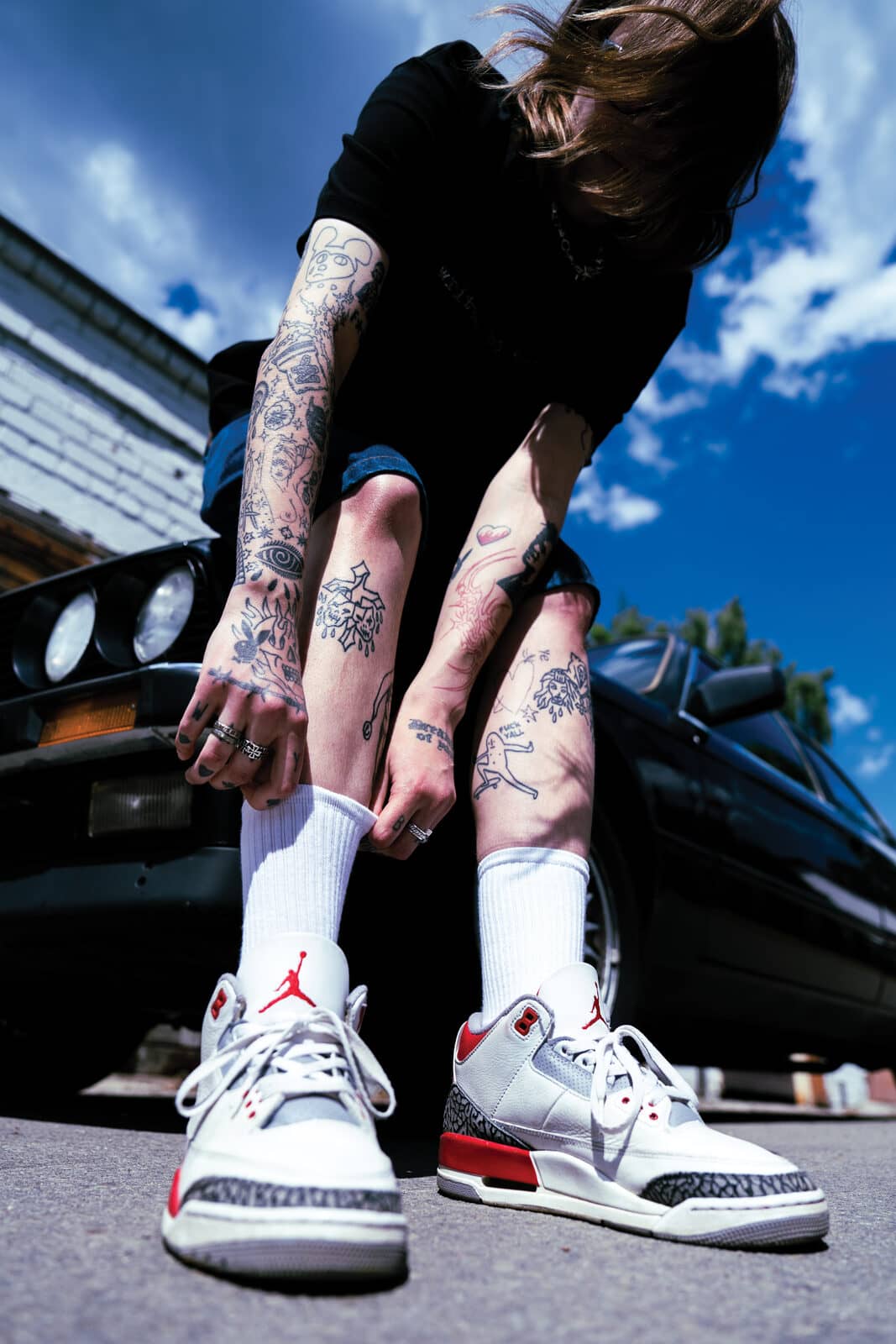 Žemas kampas prie juodo E30 – rišami Jordan 3, tatuiruotos blauzdos, slenkantis vasaros dangus.