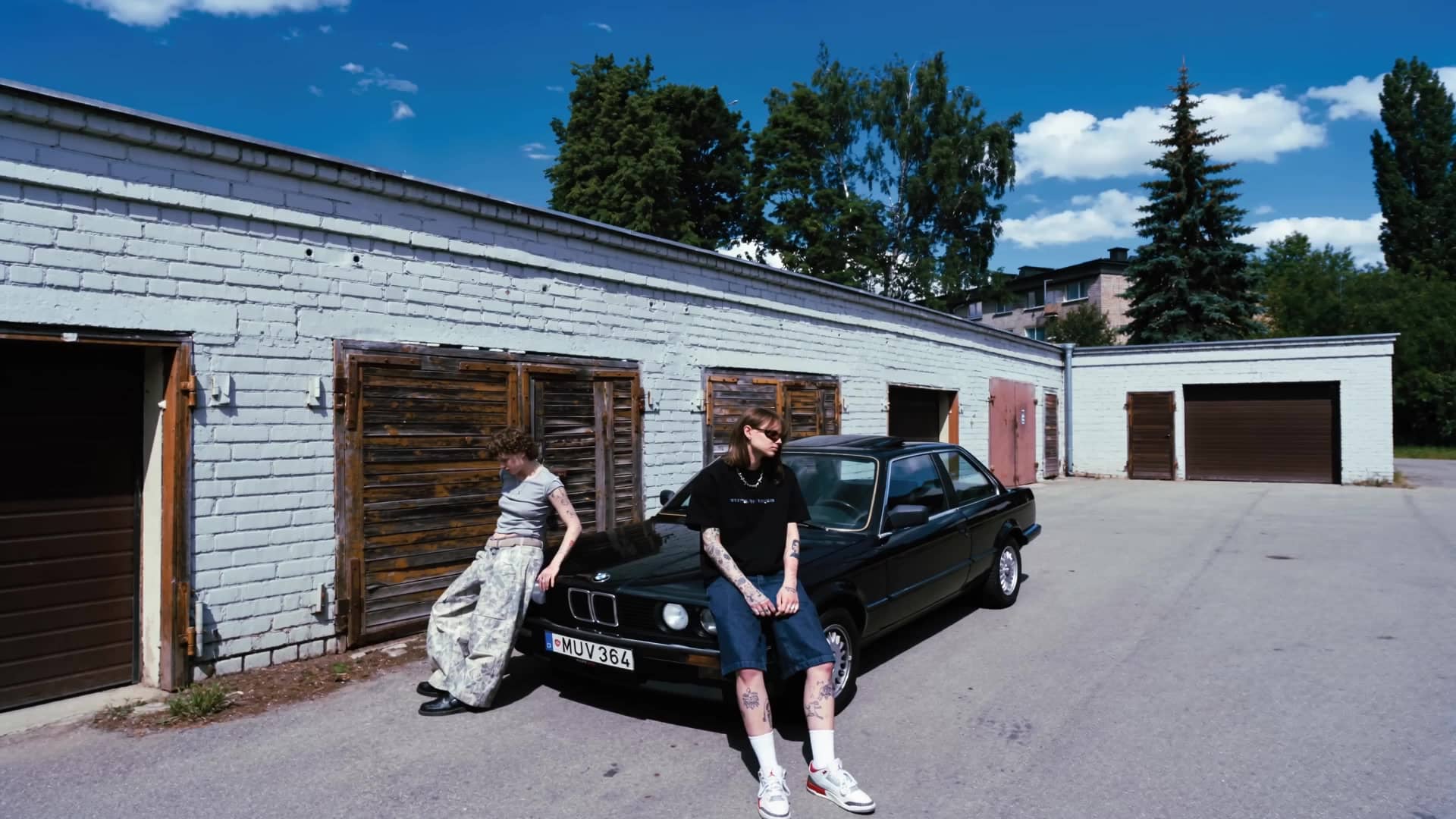 Dvi figūros ir juodas BMW E30 prie garažų – vasaros oras, slenkantys debesys.